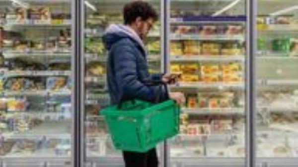 Man shopping in a supermarket while on a budget. He is looking for low prices due to inflation, standing looking at his phone in front of a row of freezers. He is living in the North East of England.