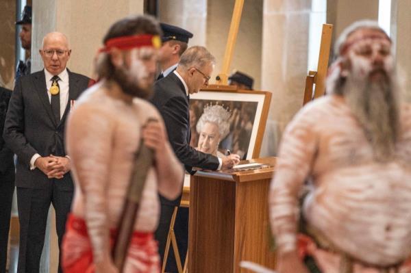 Auatralian PM Anthony Albanese signing a book in front of a picture of the queen with two Aboriginal men standing in the foreground