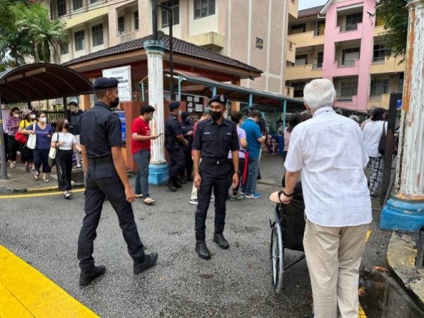 A man pushes a voter in a wheelchair into a polling station in Kuala Lumpur with police officers standing at the entrance and a queue of voters to the left