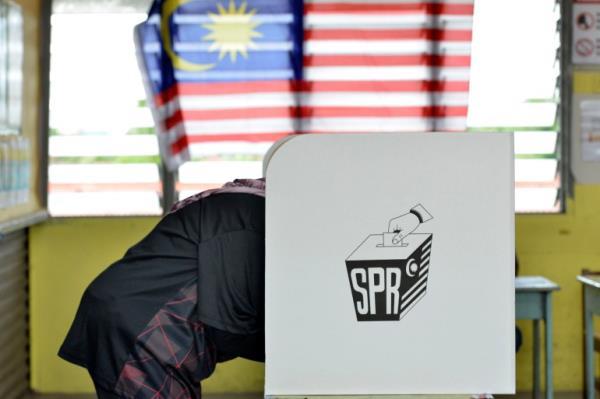 A voter hidden behind a voting booth with a Malaysian flag hanging across a window behind.