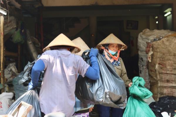 A waste collector in a conical hat walks into a collection centre with two large bags as a female collector walks out. She is also wearing a conical hat and is wearing a mask around her mouth. There are huge bags filled with waste to her left.
