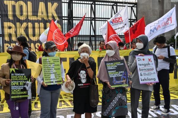A group of women outside parliament hold a protest against the new criminal code. They are masked and holding placards. Behind them are flags in red and white and a big banner saying 'TOLAK' (Reject) 