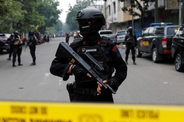 An Indonesian police officer, wearing a black BRIMOB uniform and carrying a weapon. only his eyes are visible. He is carrying a weapon and standing behind yellow police tape