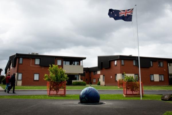Mangere Refugee Resettlement Centre in Auckland, New Zealand. It is a two storey red brick building with a brown roof and there are balconies. A New Zealand flag is flying and the garden is neat and tidy. Two people are walking towards the building carrying shopping bags