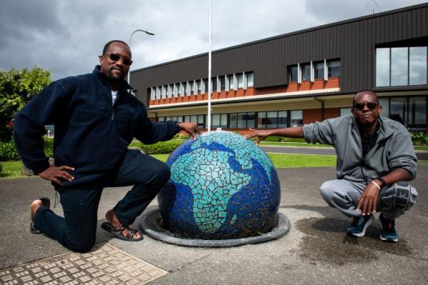 Jacques and Mahmoud squat next to a sculpture in the shape of the world outside the Mangere Refugee Resettlement Centre. They look amused